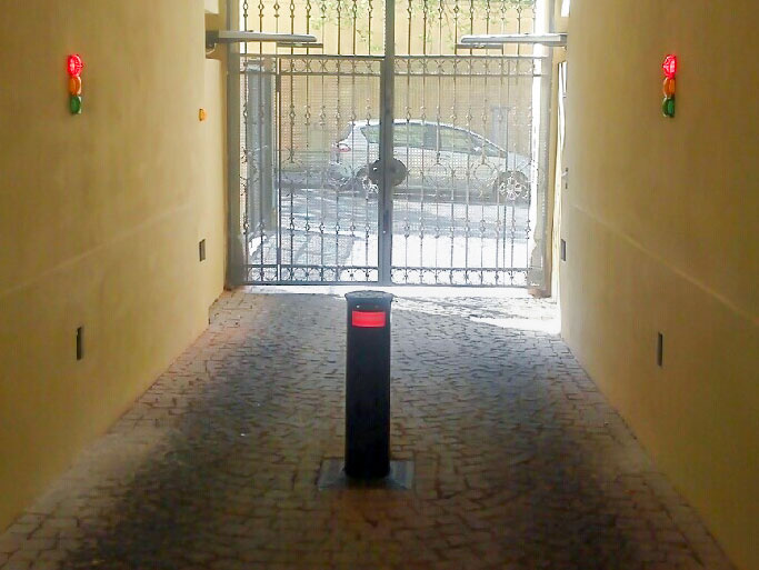 Výsuvný bezpečnostní sloup umístěný v průjezdu s červenými a zelenými signalizačními světly na stěnách a kovovou posuvnou bránou v pozadí.